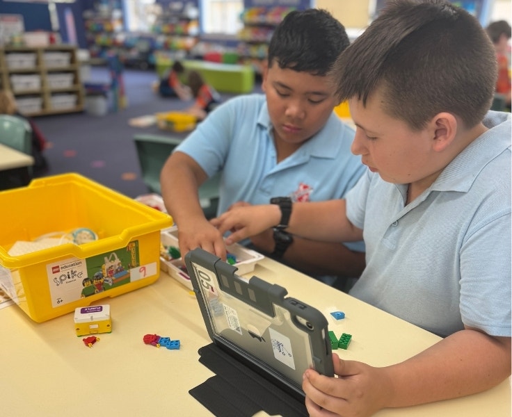 Two boys sitting at a desk working on an iPad and lego kit